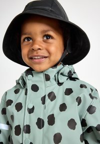 Grøn vandtæt regnjakke med sorte prikker, der har en høj krave og trykknapper. Sammen med en sort bucket hat. Glat tekstur.