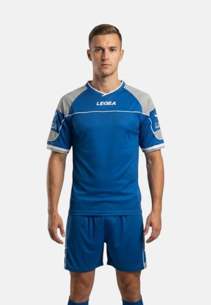 Uomo in piedi di fronte, indossa una maglia da sport blu e grigia con pantaloncini abbinati su uno sfondo bianco semplice.