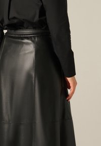 Jupe en cuir noir avec une texture lisse, présentant une taille haute, des détails de couture et une poche latérale, portée avec un haut noir.