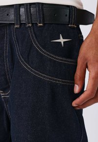 Vaqueros de mezclilla oscura con costuras doradas, que presentan un detalle metálico en forma de estrella plateada en el bolsillo delantero y un cinturón de cuero negro.