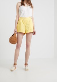 Žluté bavlněné kraťasy s předními skladbami, zapínání na dva knoflíky. Spárovány s bílým tílkem a espadrilkovými botami. Drží pletenou tašku.