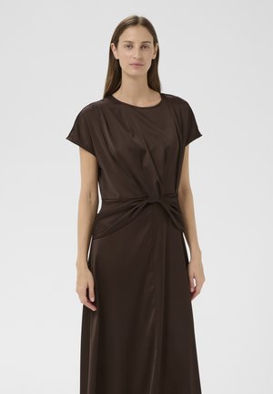 Femme aux longs cheveux bruns portant une robe en satin brun foncé à manches courtes avec un nœud torsadé sur le devant, debout devant un fond clair uni.