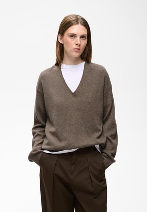 Jeune femme aux cheveux bruns et raides portant un pull marron à col en V par-dessus une chemise blanche et un pantalon marron foncé, debout les mains dans les poches.