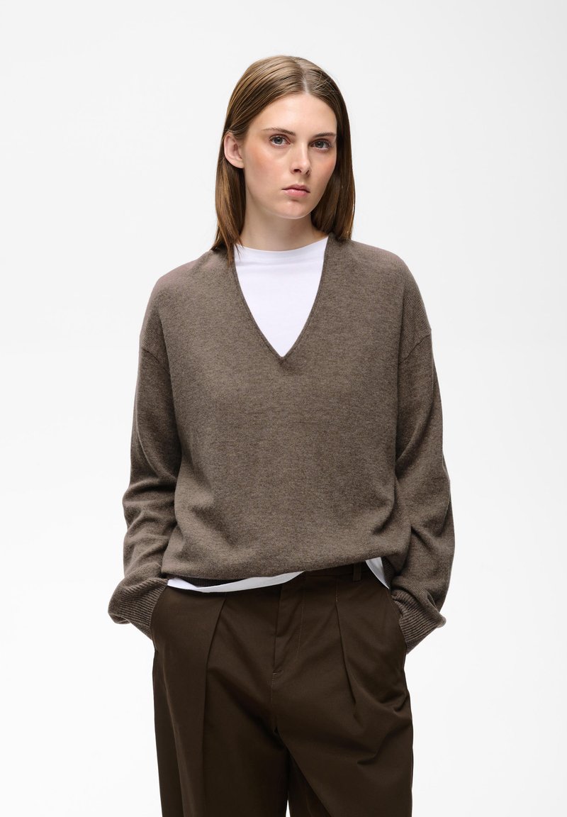 Jeune femme aux cheveux bruns et raides portant un pull marron à col en V par-dessus une chemise blanche et un pantalon marron foncé, debout les mains dans les poches.