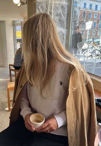 Persona ar gariem blondiem matiem, sēžot telpās, tērpusies brūnā mētelī virs baltas krekla, turot lietotu kafijas krūzi netālu no lielā loga ar skatu uz āru.