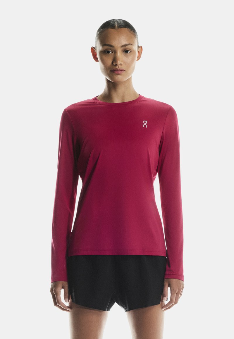 Femeie cu părul aranjat pe spate, purtând un tricou sport magenta cu mânecă lungă și pantaloni scurți negri, stând cu fața spre cameră pe un fundal alb.