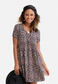 Schwarzes Kleid mit kurzen Ärmeln, V-Ausschnitt und gerafftem Taillengürtel. Mit einem pinken Herzmuster und einer fließenden Silhouette. Trägt einen schwarzen Hut.