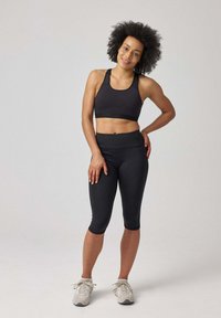 Zwarte sportieve crop top en capri leggings met een aansluitend ontwerp en een gladde textuur. Model staat met handen op de heupen, met lichte sneakers.