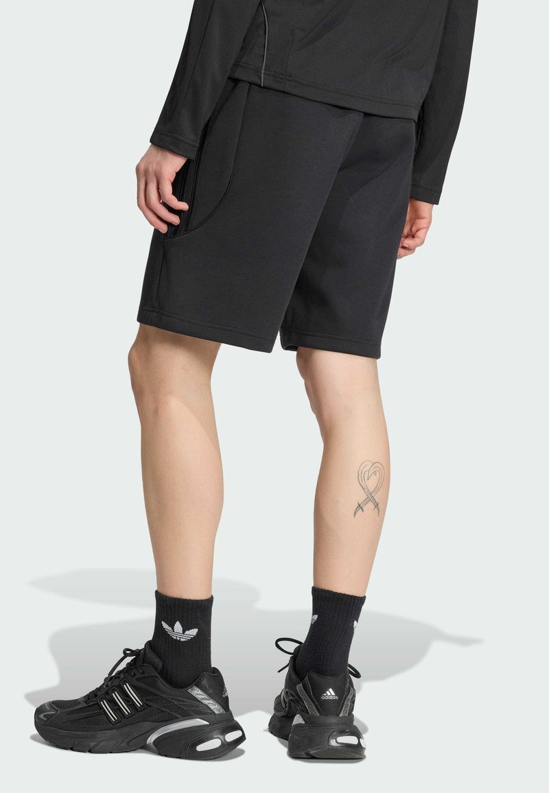 Persona che indossa pantaloncini sportivi neri, calzini Adidas neri e scarpe da ginnastica Adidas nere con un tatuaggio a forma di cuore sul polpaccio sinistro.