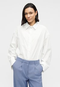 Balta pogājama krekli ar gaiši pelēkām vertikālām svītrām, kombinēta ar zilām augsta vidukļa biksēm. Garās piedurknes un brīvs siluets.