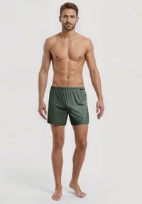 Blote man met kort bruin haar, gekleed in groene gestreepte shorts, die blootsvoets tegen een effen lichte achtergrond staat.