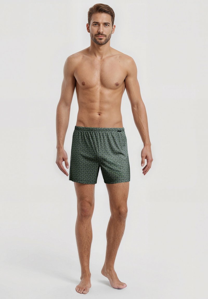 Blote man met kort bruin haar, gekleed in groene gestreepte shorts, die blootsvoets tegen een effen lichte achtergrond staat.