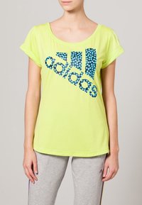 Neongul T-shirt med korta ärmar, prydd med blå prickig "adidas"-logotyp över framsidan. Mjukt tyg, avslappnad passform, enkel rund halsringning.