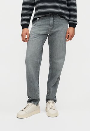 Homme portant un jean droit gris, des baskets blanches et un haut à manches longues rayé noir et gris, debout devant un fond uni.