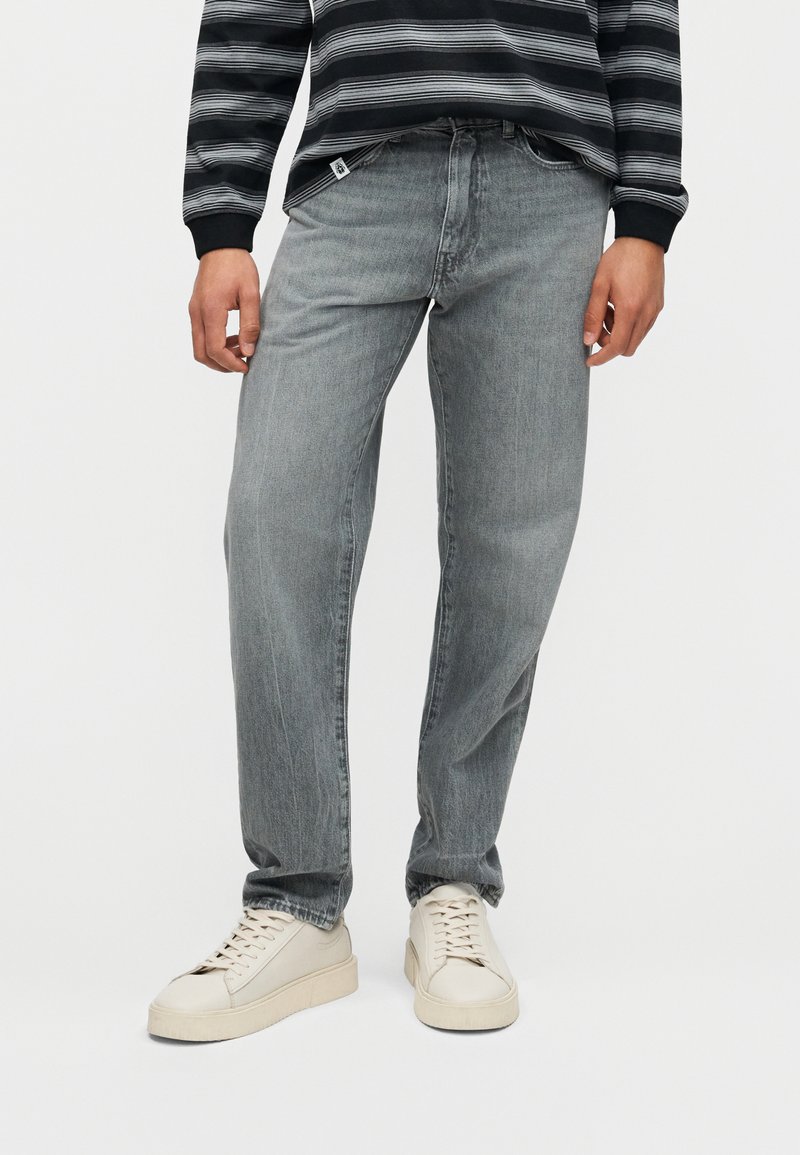 Άνδρας που φοράει γκρι τζιν με ίσια ποδάρια, λευκά αθλητικά παπούτσια και μακρυμάνικη μπλούζα με μαύρες και γκρι ρίγες, στέκεται μπροστά σε απλό φόντο.