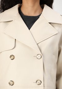 Beige läder dubbelknäppt jacka med stora knappar, skarpa slag och en slät yta, bärs över en mörk t-shirt med rundhals.