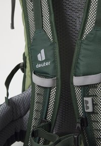 Grüner Wanderrucksack mit Netzpolsterung, grauen Trägern und dem "Deuter"-Logo. Verfügt über verstellbare Hardware und strukturierte Oberflächen.