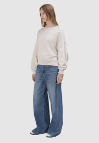 Kobieta ubrana w oversize'ową, jasnobeżową bluzę, szerokie niebieskie dżinsy i białe sportowe buty, z srebrnymi bransoletkami, stojąca na jednolitym jasnym tle.