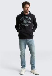 Schwarzer Kapuzenpullover mit weißem und türkisfarbenem Grafiketext, Fronttaschen, graue Innenauskleidung. Hellblaue Jeans und weiße Sneakers runden den lässigen Look ab.