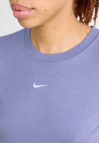 Kadar arti asmuo, dėvintis levandų spalvos Nike marškinėlius su baltu swoosh logotipu, kuris yra centre po kaklo išpjova, matoma kaklas ir apatinė veido dalis.