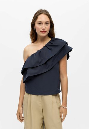 Jeune femme aux longs cheveux bruns portant un top bleu marine à épaule dénudée avec des volants et un pantalon beige à jambes larges, debout devant un fond blanc uni.