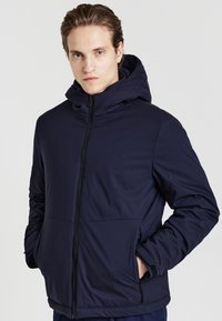 Veste isolante bleu marine avec capuche, fermeture éclair sur le devant et poches latérales. Texture lisse, design léger, présentant une coupe épurée.