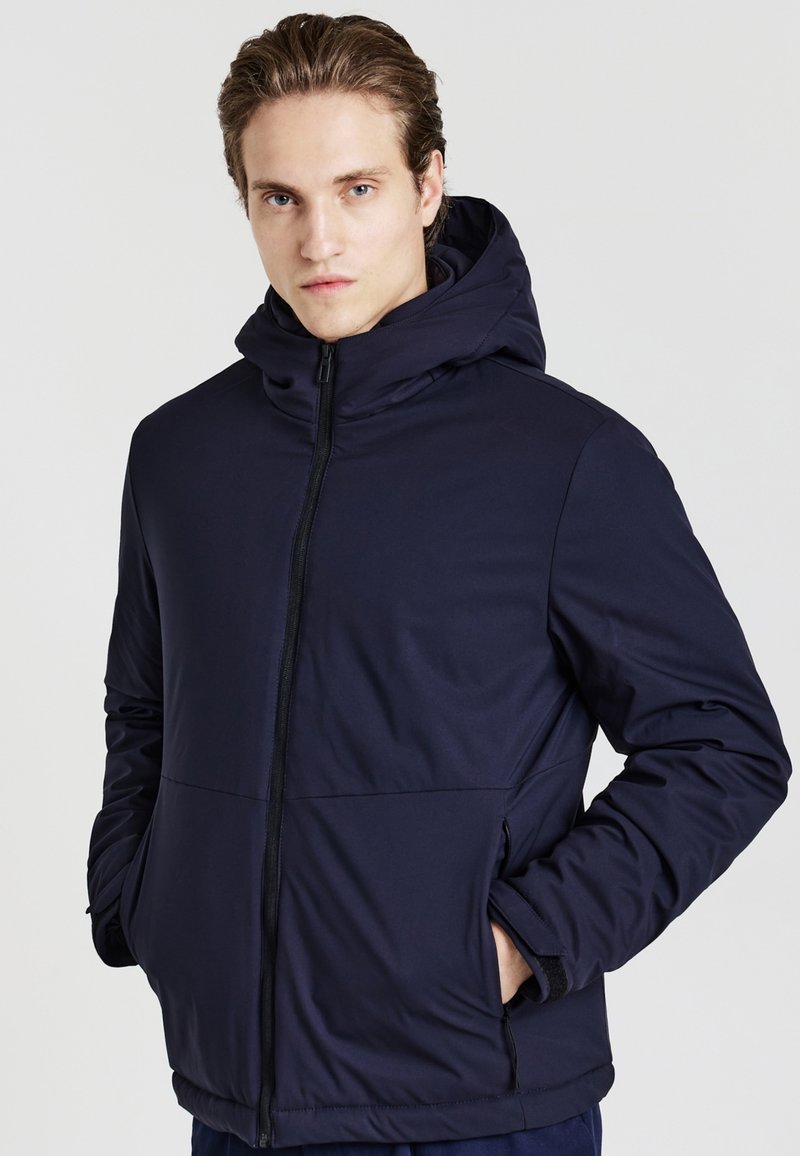 Veste isolante bleu marine avec capuche, fermeture éclair sur le devant et poches latérales. Texture lisse, design léger, présentant une coupe épurée.