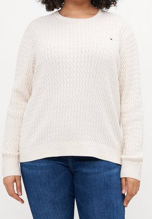 Person wearing an off-white textured long-sleeve sweater and blue jeans, shown from shoulders to hips against a white background.