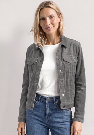 Femme blonde souriante, portant une veste grise boutonnée sur une chemise blanche et un jean bleu, debout devant un fond clair uni.