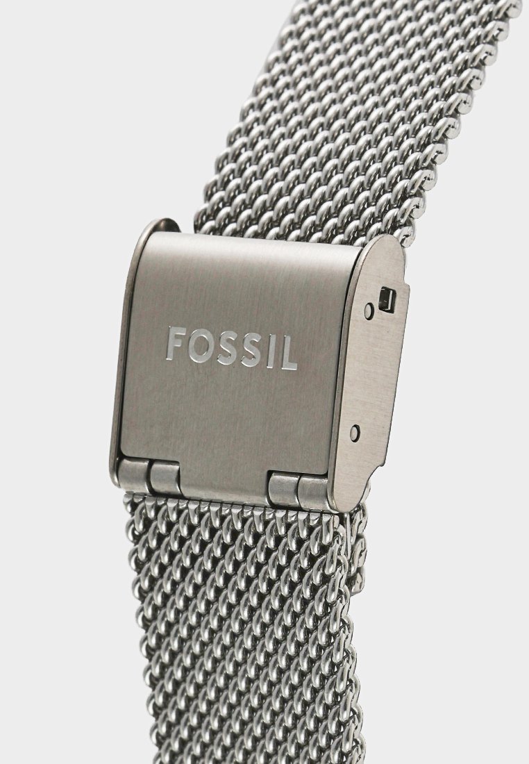 Correa de reloj de malla metálica en plata, con un cierre rectangular grabado con "FOSSIL". La correa tiene un patrón texturizado y tejido para mayor flexibilidad.