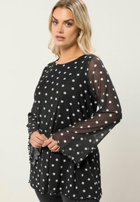 Top nero a pois bianchi con maniche lunghe trasparenti a sbuffo e orlo scallop, realizzato in tessuto leggero.