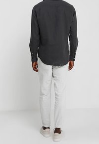 Man in een zwart T-shirt met lange mouwen, lichtgrijze joggers en witte sneakers, staand met zijn gezicht naar een witte muur.