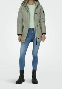 Vadderad olivgrön parkas med huva, dragkedja, dragsko i midjan, svarta detaljer, bärs över en ljusgrön tröja och blå jeans.