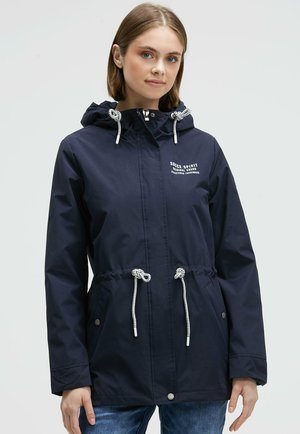 Junge Frau trägt eine marineblaue Regenjacke mit Kapuze, weißen Kordeln und dem Logo "Soccx Spirit" auf der Brust, kombiniert mit blauen Jeans.