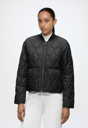 Femme portant une veste noire matelassée zippée à col rond, assortie d’un pantalon blanc, debout devant un fond uni clair.
