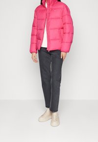 Giacca imbottita di colore rosa brillante con colletto alto e zip, caratterizzata da sezioni trapuntate, abbinata a jeans grigi e sneaker beige.