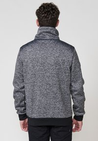 Hombre con cabello rizado que lleva una chaqueta de cremallera gris oscuro con manchas y puños y dobladillo negros, de pie de espaldas contra un fondo liso.