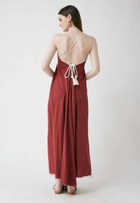 Langes, fließendes Kleid in tiefrot mit weißen Überkreuz-Trägern. Es hat ein Band und Quaste im Rücken, eine glatte Textur und ein bodenlanger Schnitt.
