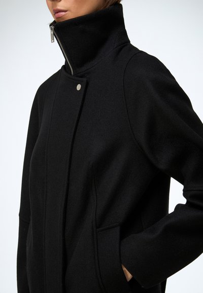 Sort jakke med høj krave, lavet af et blødt materiale. Indeholder en sølvfarvet lynlås, trykknaplukning og sidelommer. Skræddersyet fit.