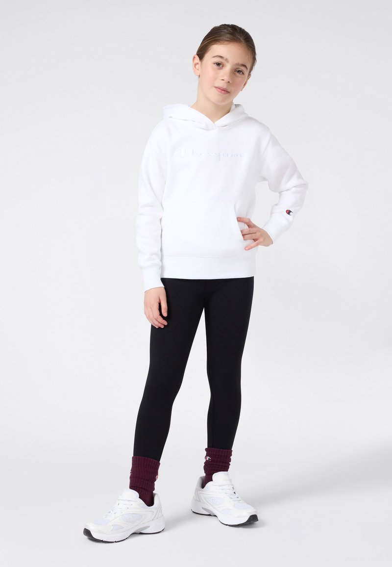 Joven de pie con la mano en la cadera, vistiendo una sudadera blanca de Champion, leggings negros, calcetines color burdeos y zapatillas blancas sobre un fondo liso.