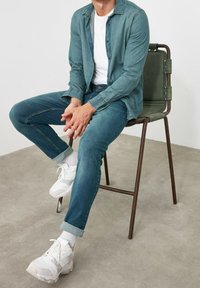 Camisa de mezclilla en azul claro, usada sobre una camiseta blanca. Jeans azul oscuro arremangados en los puños, combinados con zapatillas blancas y calcetines blancos. Sentado en una silla verde.