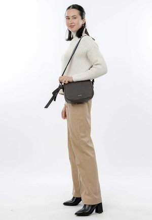 Sac bandoulière marron avec surface texturée et bandoulière réglable, associé à un pull en tricot crème et un pantalon beige, porté avec des bottes à talons noires.