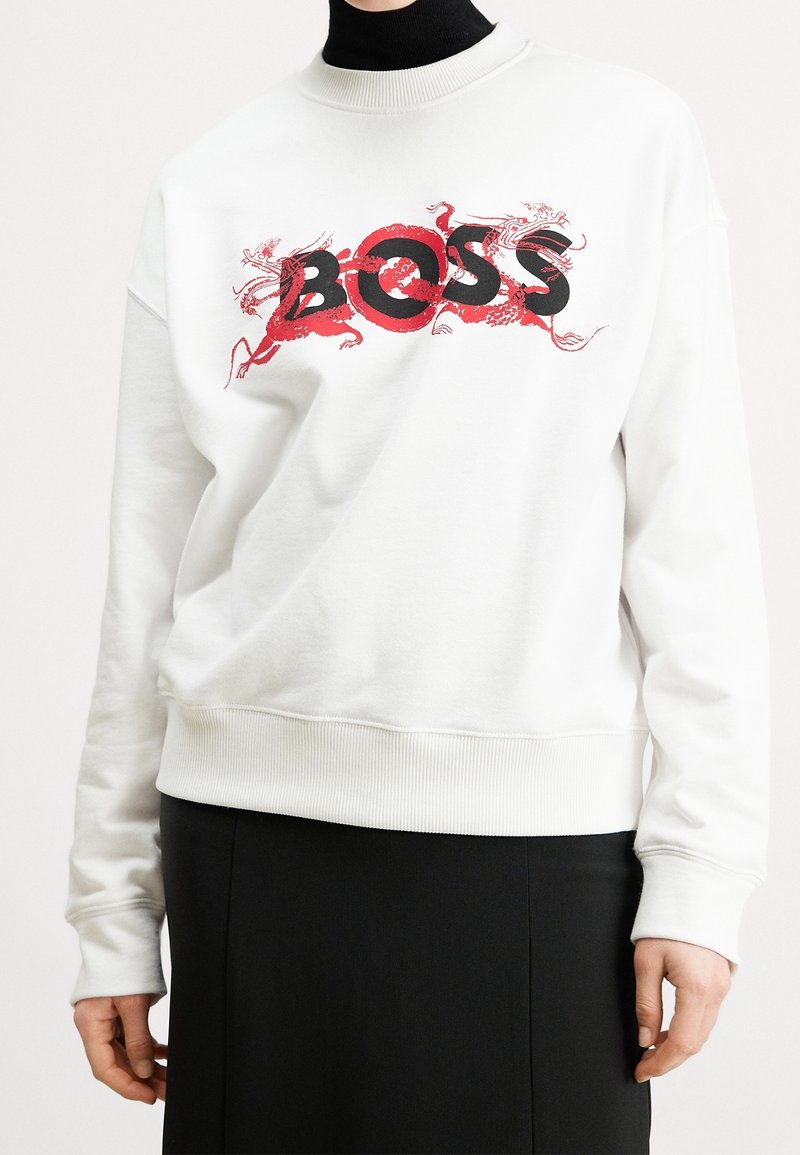 Sweat blanc avec texte "BOSS" noir et rouge et motif dragon rouge porté sur un col roulé noir et une jupe noire.