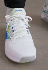 Shoe atlético branco com parte superior em malha, acentos verdes e azuis, atacadores planos e sola texturizada. Apresenta o logótipo da Reebok na língua.