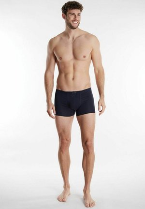 Lächelnder, oberkörperfreier Mann steht barfuß und trägt schwarze Boxer-Shorts vor einem schlichten weißen Hintergrund.