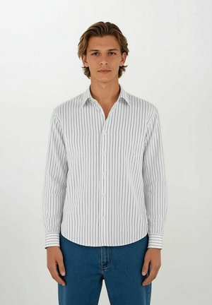 Ung man med medellångt hår som bär en vit, långärmad skjorta med vertikala ränder och blå jeans, stående mot en enfärgad vit bakgrund.