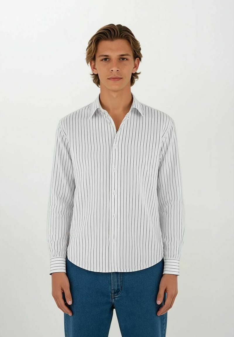 Ung mann med middels langt hår iført en hvit, langermet skjorte med vertikale striper og blå jeans, som står mot en ensfarget hvit bakgrunn.