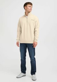 Beige zip-up sweatshirt met ribgebreide mouwen en zoom, voorzien van een zwart "A.R.C." logo op de linkerkant van de borst, gecombineerd met donkere jeans en grijze sneakers.