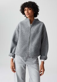 Giacca grigia con zip realizzata in tessuto testurizzato, con spalle scese e polsini arrotolati. Abbinata a pantaloni in denim grigio chiaro.