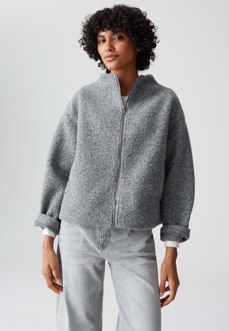 Giacca grigia con zip realizzata in tessuto testurizzato, con spalle scese e polsini arrotolati. Abbinata a pantaloni in denim grigio chiaro.